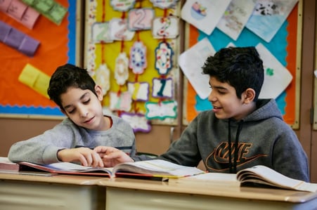 Two students point to a page of a book spread across their desks. 