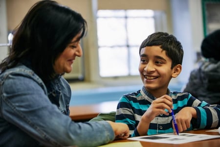 An elementary student points at a paper on the table and smiles at their family partner. 